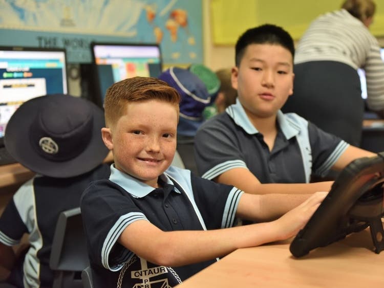 two students sitting at a desk working on an ipad with more students working on computers in the backgroud