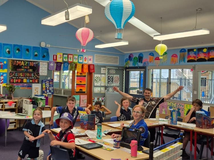 small class of students sitting at their desks with their teachers. the room has many colourful posters and there are floating balloons on the ceiling